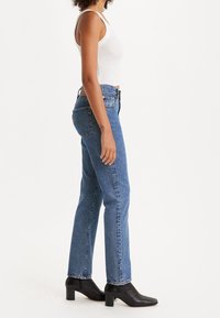 Jeans de mezclilla azul con un corte recto, cintura alta y costuras en contraste. Combinados con una camiseta sin mangas blanca y botas de tacón negro hasta el tobillo.