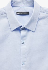 Camisa de manga corta azul claro hecha de tejido texturizado. Presenta un cuello clásico, dos botones azul claro y un diseño de corte ajustado.