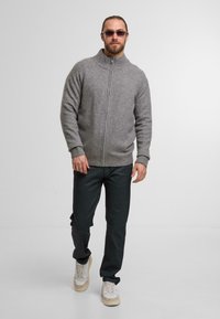 Szara rozpinana kardigan z wysokim kołnierzem, zestawiona z ciemnymi spodniami i jasnymi sneakersami. Model stoi na tle jednolitego białego tła.