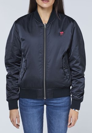 Schwarze Bomberjacke mit einem Frontreißverschluss, Rippbündchen und Saum. Verfügt über Seitentaschen und ein kleines rotes Logo auf der linken Brustseite.