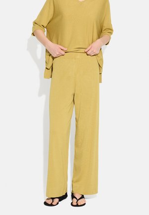 Osoba ubrana w luźne musztardowożółte spodnie i pasującą górę, trzymająca pas, oraz czarne sandały z odkrytymi palcami na białym tle.