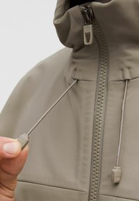 Chaqueta de cremallera gris claro con cuello alto, que cuenta con un ajustador de cordón y una textura de tela suave. Cremallera frontal y acento de toggles.