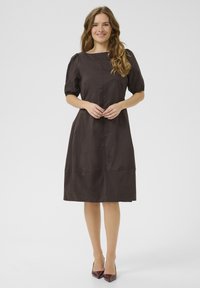 Rjava obleka s kratkimi napihnjenimi rokavi, okroglim izrezom in dolžino do kolen. Narejena je iz gladkega materiala s sprednjim šivom in minimalnimi detajli.