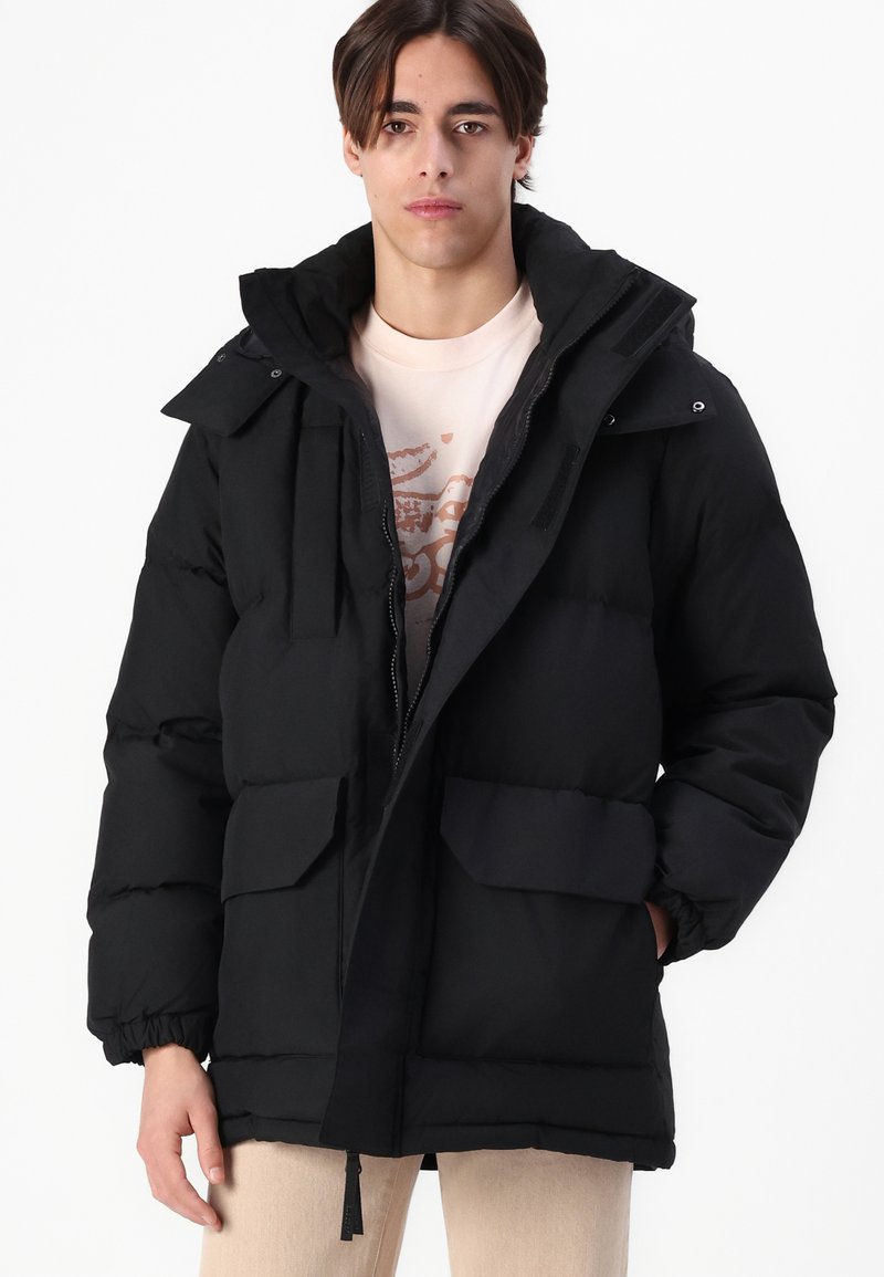 Veste matelassée noire avec un col haut, une fermeture éclair et deux poches plaquées à l'avant. Fabriquée en tissu lisse avec une texture douce.