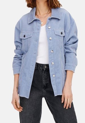 Kvinde iført en lyseblå kordfløjlsjakke med knapper, hvid skjorte, sorte højtaljede jeans og lagdelte guldkæder.