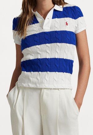 Femme portant un polo en maille torsadée rayé bleu et blanc à manches courtes et un pantalon blanc plissé, mains dans les poches.
