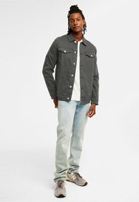Grå denimjacka med krage, två bröstfickor och silverfärgade knappar. Bärs över en vit skjorta, i kombination med ljusblå jeans och grå sneakers.