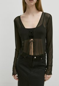 Schwarzes, transparentes Langarmoberteil mit tiefem V-Ausschnitt und frontalem Bindedetail, über einem schwarzen, figurbetonten Tanktop getragen, kombiniert mit einem Rock aus dunklem Denim.