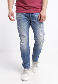 Hombre vestido con jeans rotos de color azul deslavado, zapatillas blancas y una camisa roja, caminando contra un fondo blanco liso.