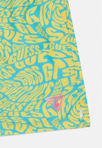 Costumi da bagno da uomo con uno sfondo blu e testo ripetuto in giallo "GUESS", e un logo triangolare rosa con un accento di palma.
