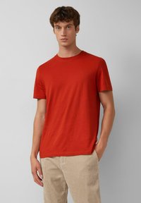 Kurzärmliges rotes T-Shirt aus weicher Baumwolle, mit rundem Ausschnitt und einem dezenten Logo auf der Brust, kombiniert mit beigen Cordhosen.