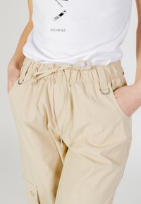 Pantaloni beige con vita regolabile, polsini elasticizzati e tasche laterali. Tessuto liscio e leggero con dettagli metallici vicino alle tasche.