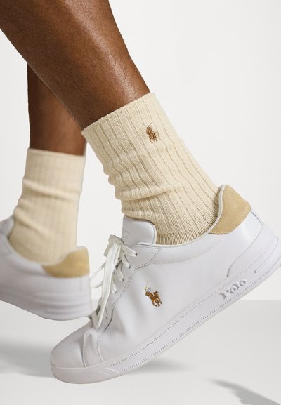 Jalad kreemist soonikute ja tikitud logo ning valgete paeltega tossudega, tõstetud valge tausta vastu.