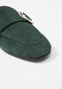 Chaussure en daim verte avec un bout pointu, présentant une large sangle et un accent de boucle en argent. Texture lisse et semelle plate noire.