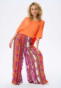 Pomarańczowa, oversized koszulka zestawiona z długimi, wzorzystymi spodniami w różowe, fioletowe i pomarańczowe tribalne wzory z paskami i kształtami geometrycznymi.