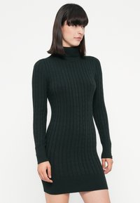 Mujer luciendo un vestido suéter de punto trenzado negro ajustado con cuello alto y mangas largas, contra un fondo claro y liso.