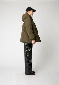 Pufferjacke in olivgrün mit Kapuze, Seitentaschen und gestepptem Design. Kombiniert mit hochglänzenden schwarzen Vinylhosen und einer schwarzen Mütze.