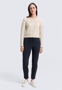 Jasnobeżowy prążkowany sweter, ciemnoniebieskie spodnie ze ściągaczem oraz białe trampki, noszone przez stojącą modelkę na jednolitym tle.