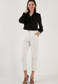 Black blouse with a tied neckline, long sleeves, and a relaxed fit. White pants with a straight cut, side pockets, and hemmed ankles. Black heeled sandals.