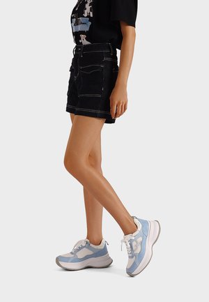 Donna in piedi con le gambe incrociate, indossa pantaloncini neri, una maglietta nera con stampa di cane e sneakers chunky bianche e blu su sfondo bianco.