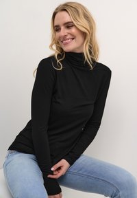 Melnas garās piedurknes turtleneck krekls mīkstā audumā ar pieguļošu dizainu. Modelis valkā gaiši zilos džinsus, sēžot uz balta fona.