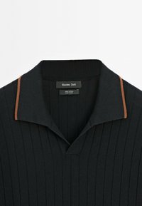Polo shirt in cotone nero con texture a coste, scollo a V e un contrasto marrone sul colletto. Etichetta del brand visibile all'interno.