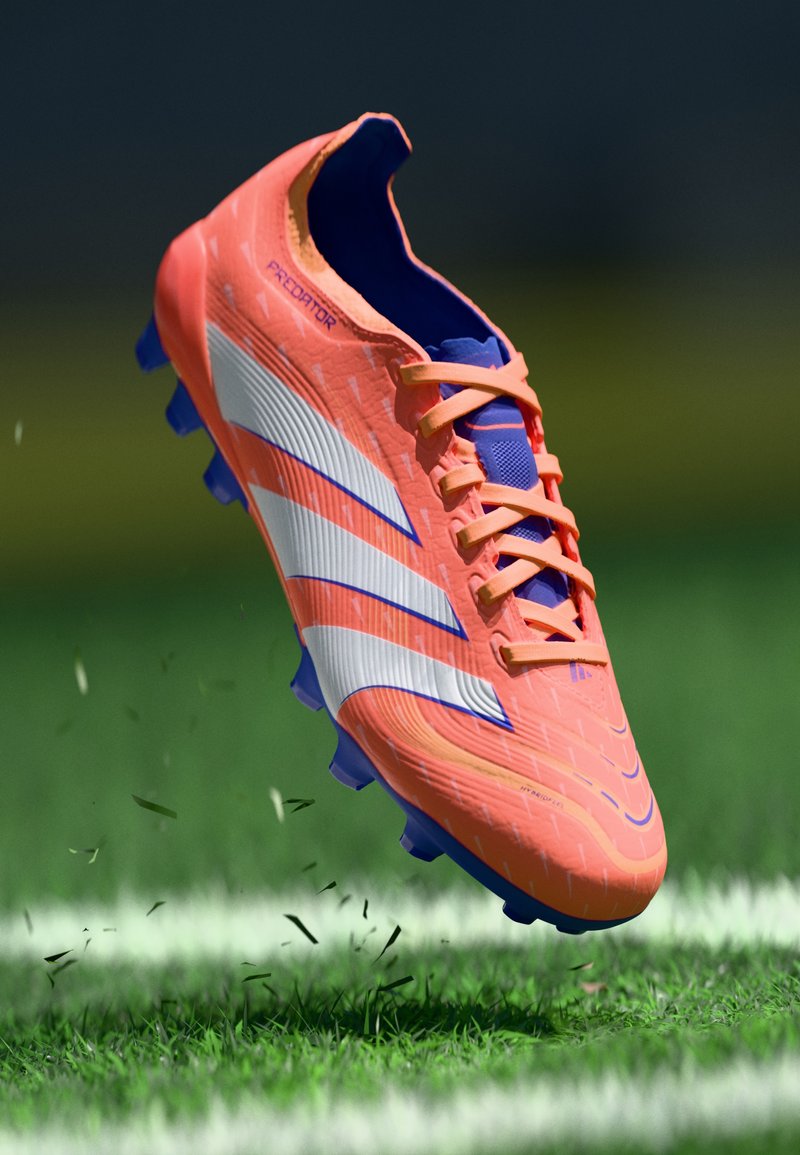 Scarpe da calcio arancioni e blu con tomaia testurizzata, strisce bianche e blu e tacchetti viola, sospese sopra un prato verde.