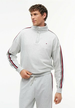 Hombre con chándal gris claro con rayas rojas, blancas y azules en las mangas y los pantalones, de pie con una mano cerca de la cintura.