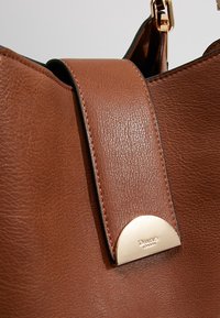 Brown leather handbag with a smooth texture, featuring a wide strap and a gold-tone metal accent. The edge has black trimming.