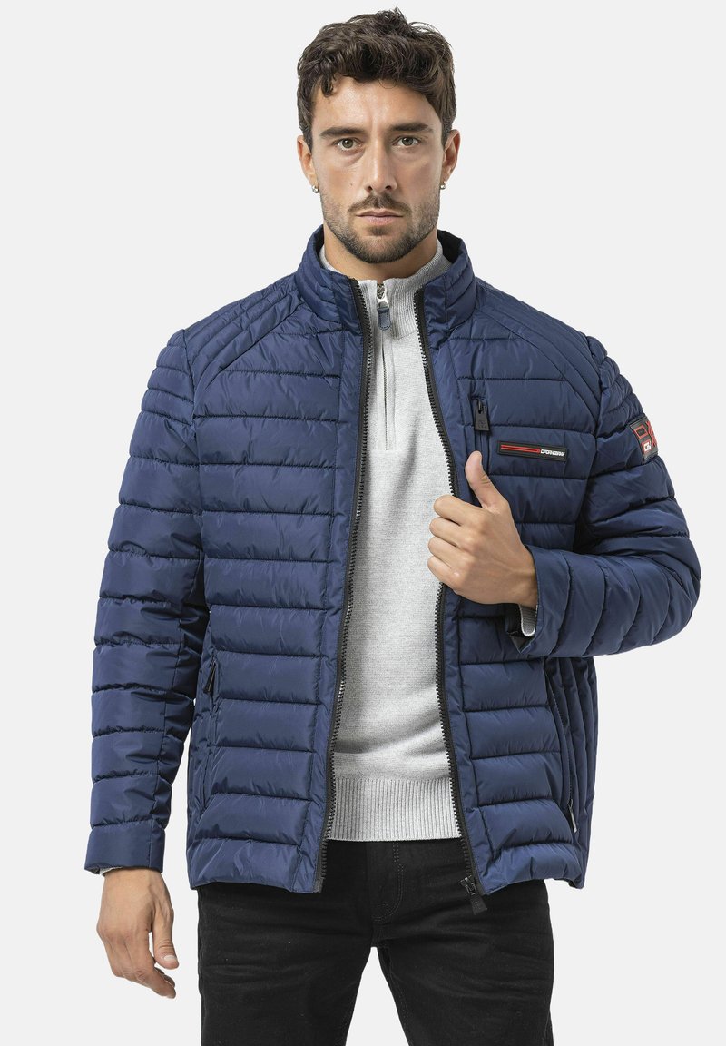 Doudoune matelassée bleu marine avec col montant, fermeture éclair à l'avant et poches latérales. Dotée de bandes rouges en accents et de détails avec logo.