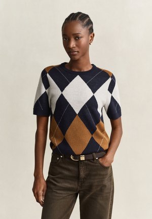 Femme aux cheveux tressés portant un pull à manches courtes à motif losanges bleu marine, blanc et marron, associé à un pantalon foncé et une ceinture, debout avec une main dans la poche.