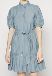 Světlemodré denimové šaty s límečkem, nafouknutými rukávy, zapínáním na knoflíky, páskem v pase a vrstveným volánkovým lemem. Hladká textura a volný střih.