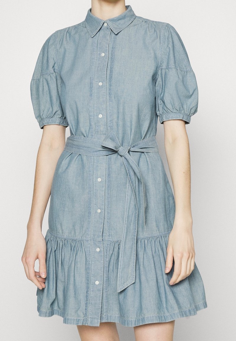 Světlemodré denimové šaty s límečkem, nafouknutými rukávy, zapínáním na knoflíky, páskem v pase a vrstveným volánkovým lemem. Hladká textura a volný střih.