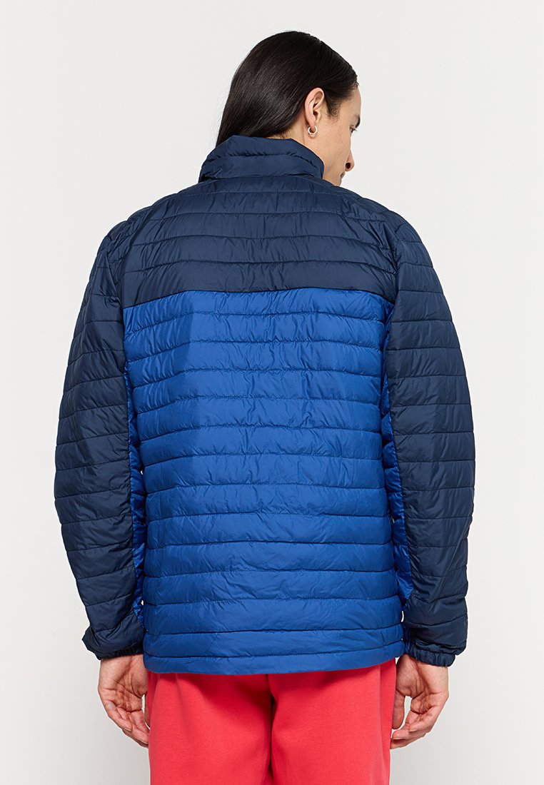 Hombre con cabello largo y oscuro, vestido con una chaqueta de dos tonos en azul acolchada y pantalones rojos, de espaldas frente a un fondo claro.