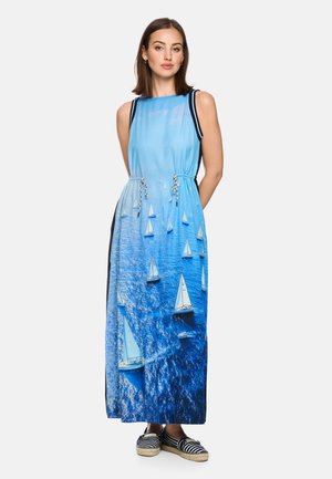 Frau trägt ein ärmelloses blaues Maxikleid mit weißem Segelboot-auf-Ozeanwellen-Muster, kombiniert mit schwarz-weiß gestreiften Espadrilles.