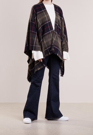 Osoba oblečená v tmavém kostkovaném ponču přes bílý svetr, tmavé rozšířené džíny a bílé tenisky stojí na světlé podlaze.