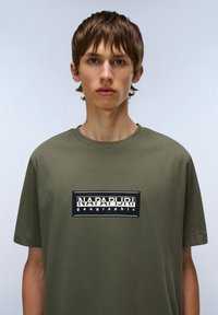 Oliven grøn kortærmet t-shirt lavet af bomuld med et sort rektangulært logo, der har "NAPAPURRI geographic" trykt centralt.