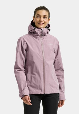 Lächelnde Frau in einer hellvioletten Kapuzenjacke mit Reißverschluss und schwarzen Hosen, steht vor einem hellen, schlichten Hintergrund.