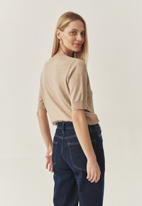 Beige kötött felső rövid ujjakkal, kerek nyakkivágással, sima textúrával, sötét kék farmernadrággal párosítva, amelyen látható varrás és hátsó zsebek találhatóak.