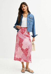Giacca di jeans sopra una canotta bianca, abbinata a una lunga gonna rosa con motivi floreali rossi. La modella tiene una piccola borsa beige.