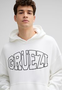 Witte hooded sweatshirt met het woord "GRÜEZI" in vetgedrukte zwarte letters. Gemaakt van een gestructureerde stof met een ontspannen pasvorm en een trekkoordkap.
