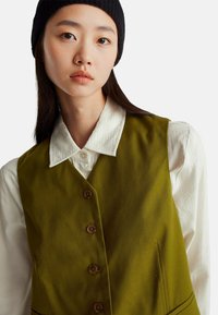 Gilet verde oliva con una texture liscia, caratterizzato da cinque bottoni marroni, indossato sopra una camicia bianca con un delicato motivo testurizzato e un colletto rotondo.