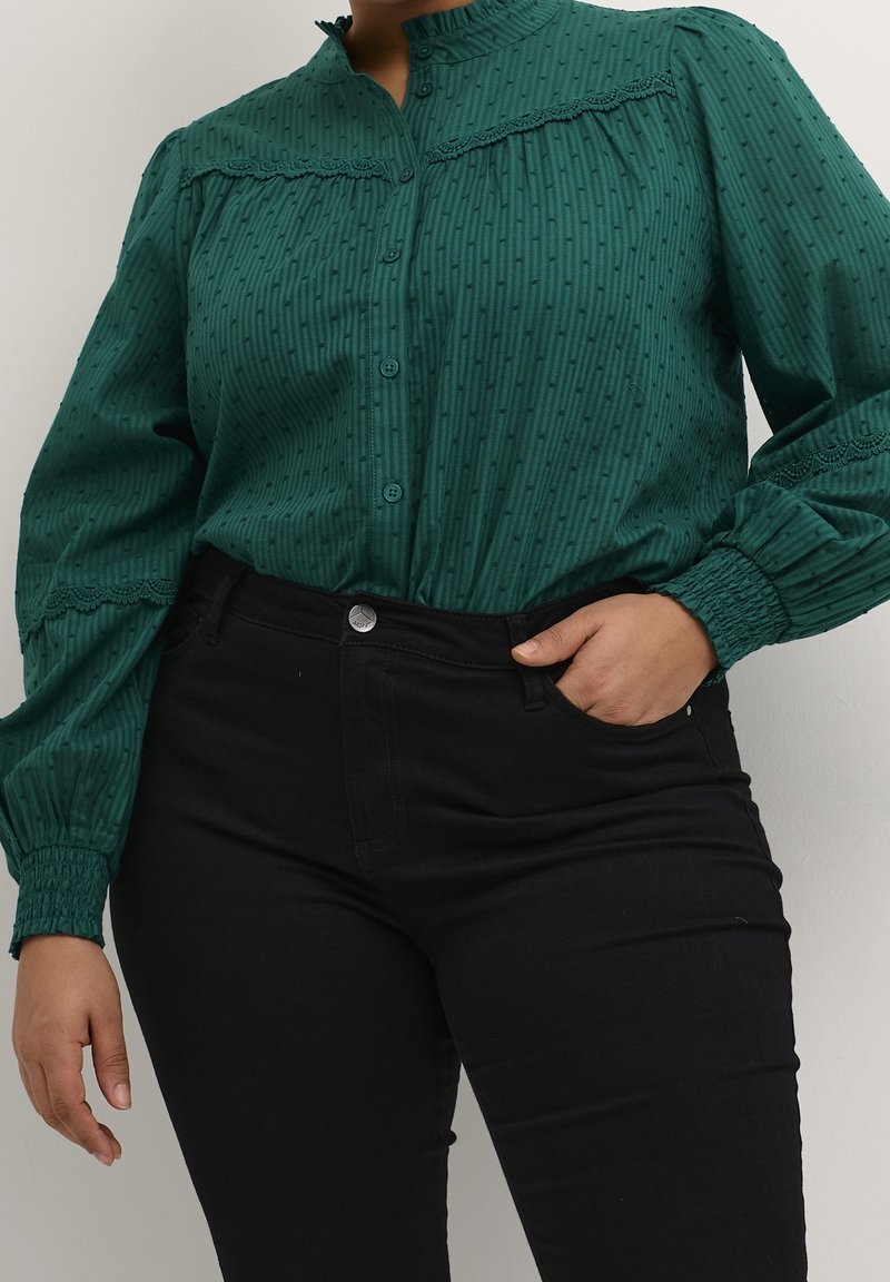 Groene blouse met knopen, voorzien van een gestructureerd patroon, hoge kraag, gathered mouwen en kantafwerking. Gecombineerd met zwarte strakke broek.