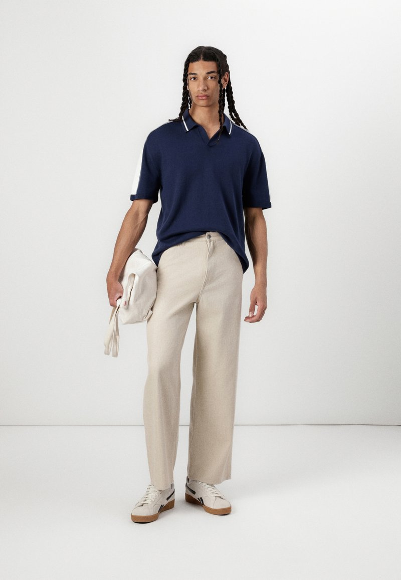 Granatowa koszulka polo z krótkim rękawem i białymi akcentami, beżowe spodnie z szerokimi nogawkami, trzymając kremową torbę, nosząc beżowe trampki z brązowymi podeszwami.