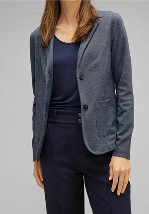 Vrouw met een aangesloten grijze blazer over een marineblauwe top en hooggesneden marineblauwe broek, staand tegen een effen grijze achtergrond.