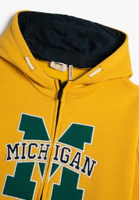 Felpa gialla con zip, caratterizzata da una "M" verde e la scritta "MICHIGAN" in nero, con cappuccio foderato in blu navy e accenti bianchi sulla zip.