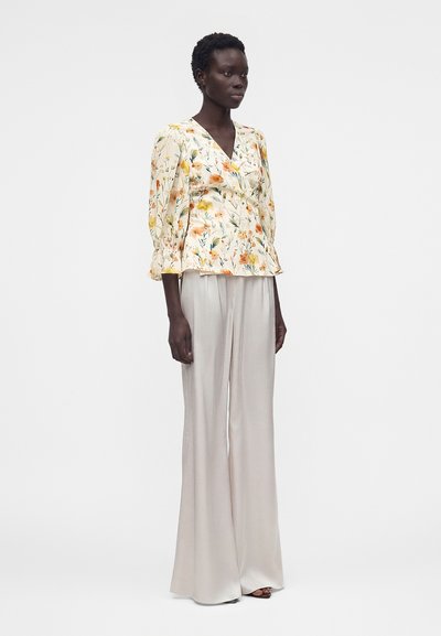 Γυναίκα στέκεται φορώντας μια κρεμίστικη μακρυμάνικη μπλούζα με λουλούδια και λευκό φαρδύ σατέν παντελόνι.