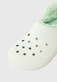 Weiße Clogs ohne Schnürsenkel mit runden Lüftungsöffnungen und einem hellgrünen, flauschigen Innenfutter, das an der Ferse sichtbar ist.