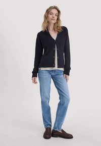 Vrouw die staat en een zwarte knopen cardigan, een beige top, lichtblauwe jeans en donkerbruine instappers draagt, tegen een effen lichte achtergrond.