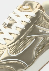 Αθλητικό παπούτσι σε μεταλλικό χρυσό χρώμα με υφή από πλεκτό πλέγμα, λευκά κορδόνια και εμφανή λογότυπα. Η σόλα είναι λευκή με καφέ πέλμα.